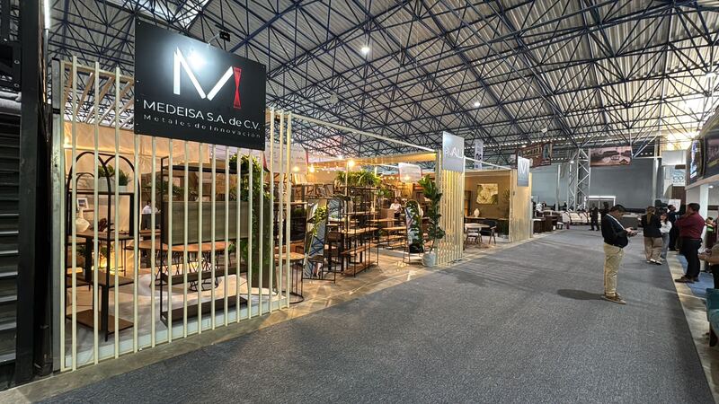 Stand de MEDEISA en Expo Muebles Ocotlán: letrero negro con el logo, celosía de listones claros y libreros, mesas y estanterías de acero negro y madera iluminados con luz cálida